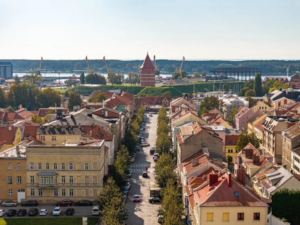 Panoraminis Klaipėdos senamiesčio vaizdas su raudonų čerpių stogais, centrine gatve ir uosto kranais tolumoje.