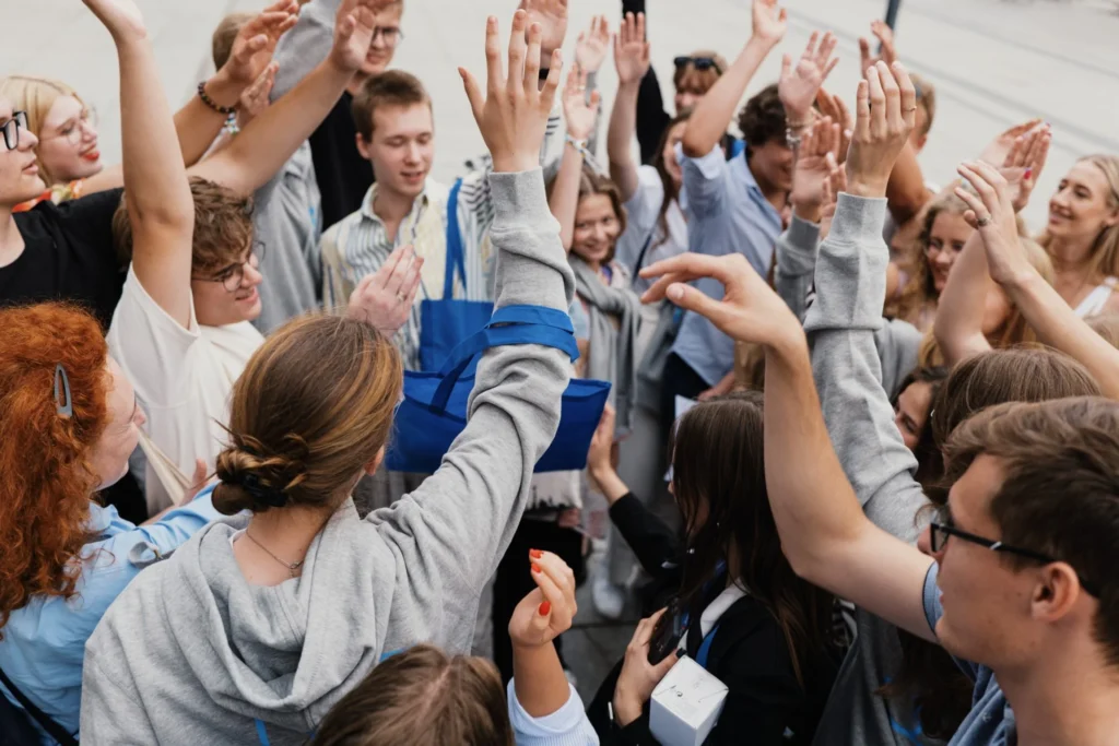 Grupė jaunų žmonių, sustojusių ratu lauke, džiaugsmingai kelia rankas į viršų.
