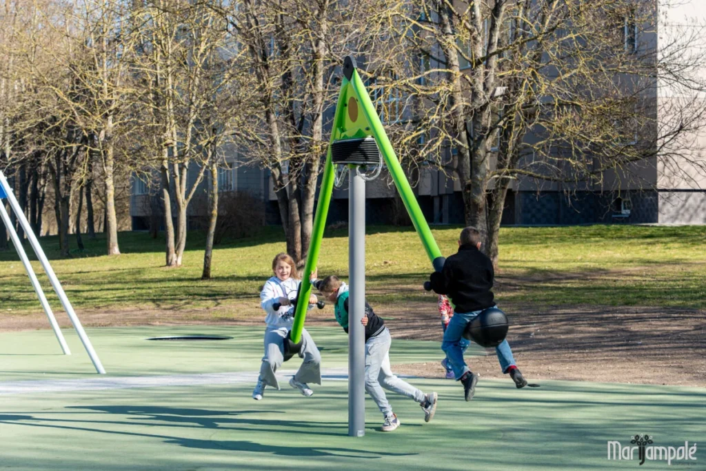 Vaikai džiaugsmingai supasi ant žalios modernios karuselės miesto aikštelėje saulėtą pavasario ar rudens dieną.