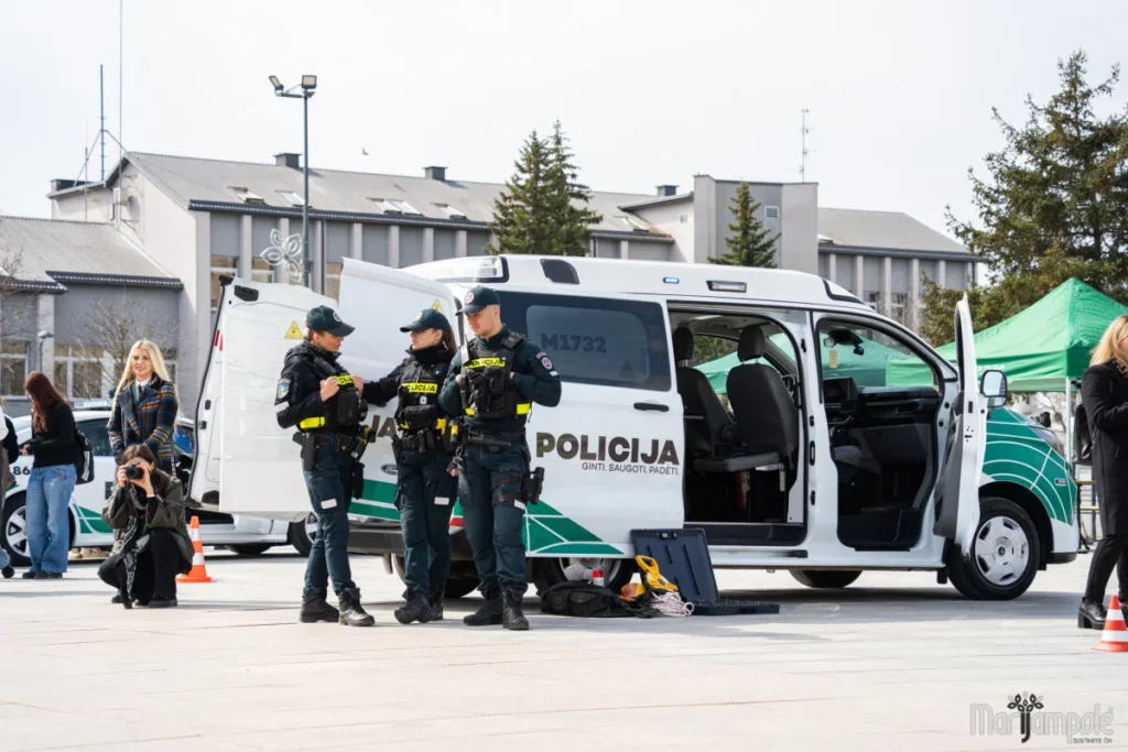Trys Lietuvos policijos pareigūnai stovi šalia balto policijos mikroautobuso atviromis durimis aikštėje dienos metu.