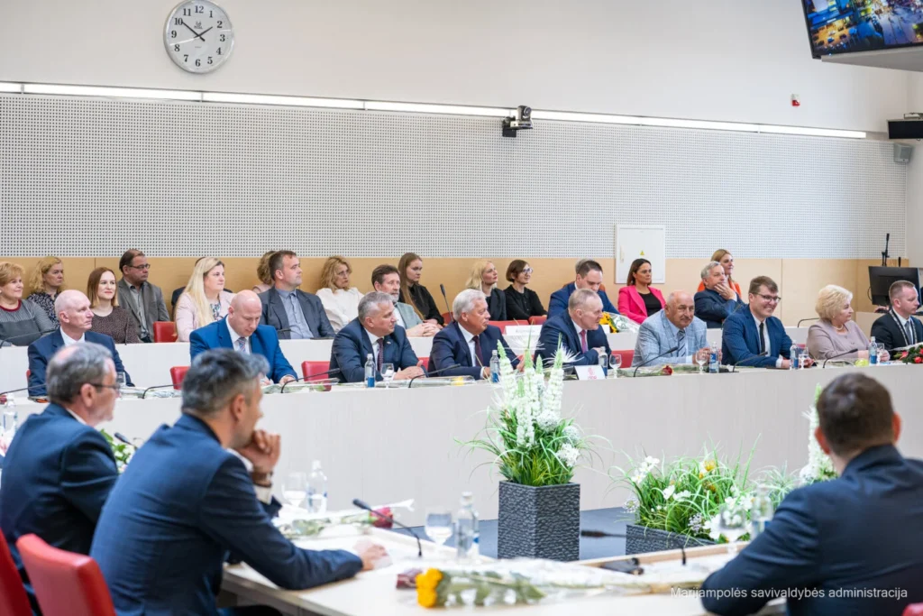 Kostiumuoti asmenys sėdi prie ilgų stalų modernioje posėdžių salėje su gėlėmis ant stalo.