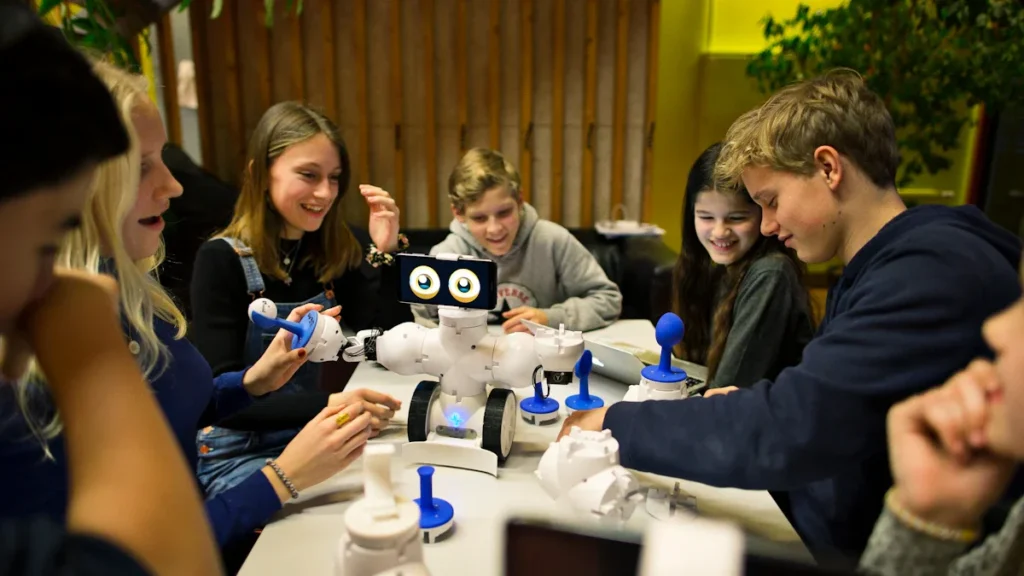 Grupė mokinių susidomėję stebi ir valdo baltą edukacinį robotą modernioje mokyklos klasėje.
