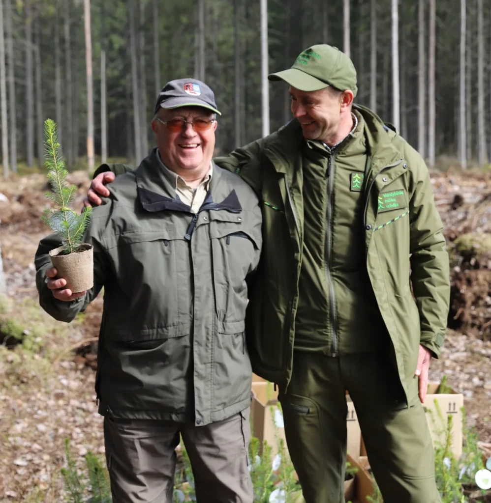 Du vyrai miške: vienas laiko eglės sodinuką, kitas vilki miškininko uniformą ir šypsosi.