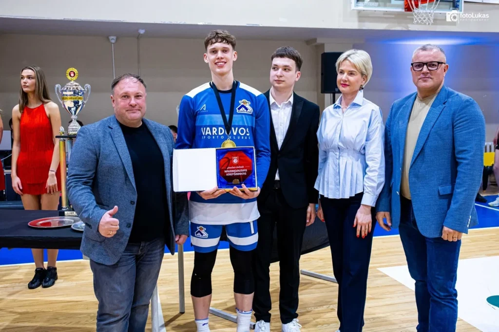 Jaunas krepšininkas mėlyna apranga laiko apdovanojimą, stovėdamas šalia keturių oficialių asmenų sporto salėje.