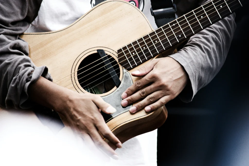 Artimas vaizdas į muzikanto rankas, laikančias šviesaus medžio akustinę gitarą su metalinėmis stygomis.