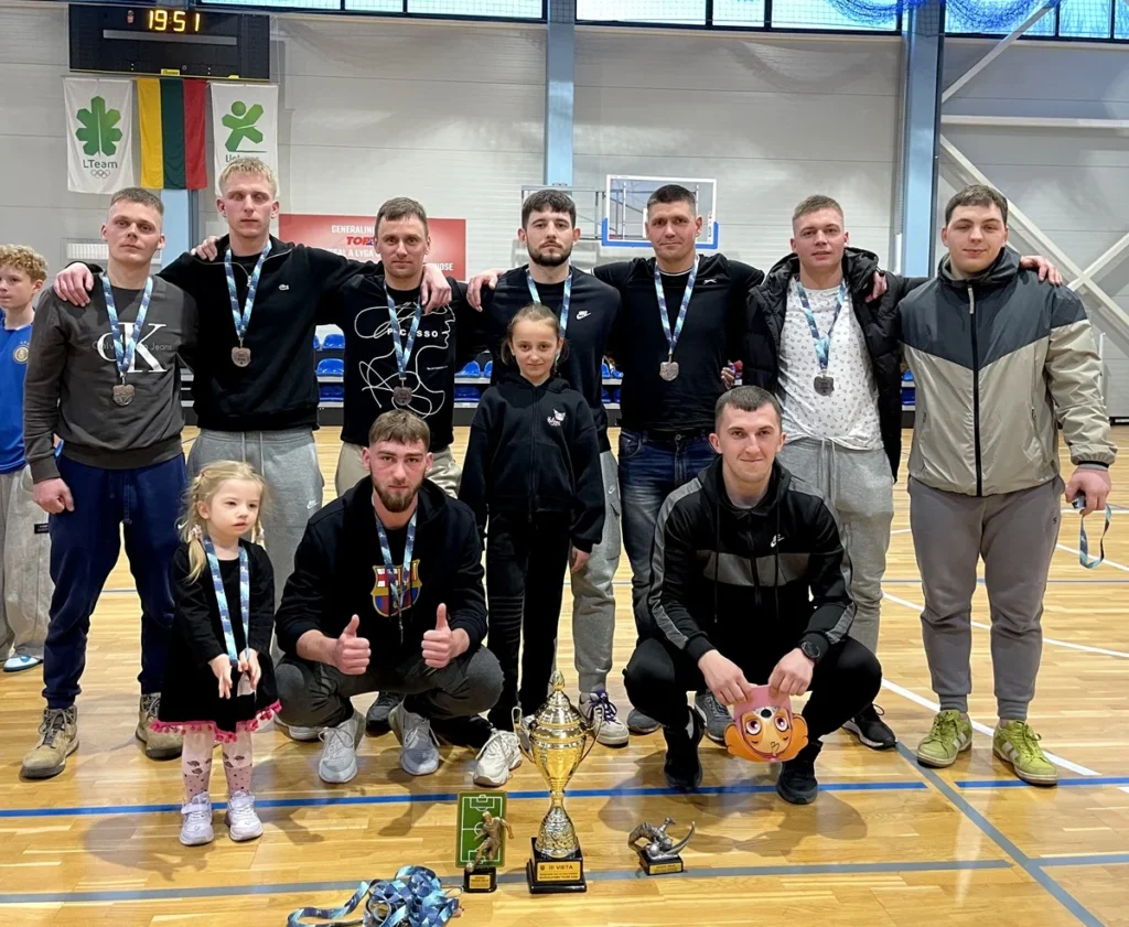 Grupė vyrų su medaliais ir vaikai pozuoja sporto salėje prie didelės auksinės taurės bei trofėjų.