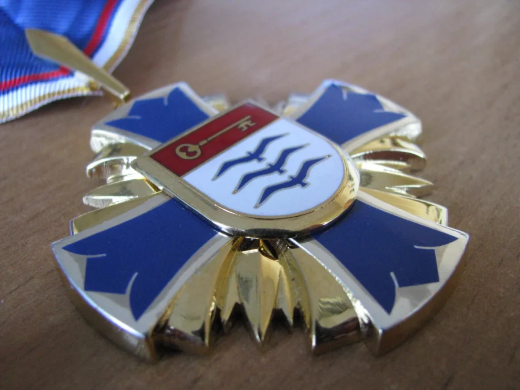 Auksinis kryžiaus formos apdovanojimas su mėlyna emale ir Palangos miesto herbu centre ant medinio paviršiaus.