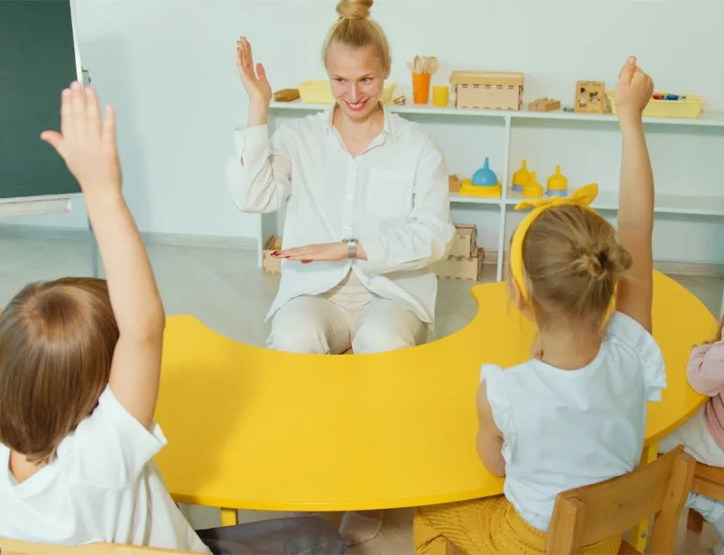 Specialistė gestų kalba bendrauja su mokiniais prie geltono pusapvalio stalo šviesioje edukacinėje erdvėje.