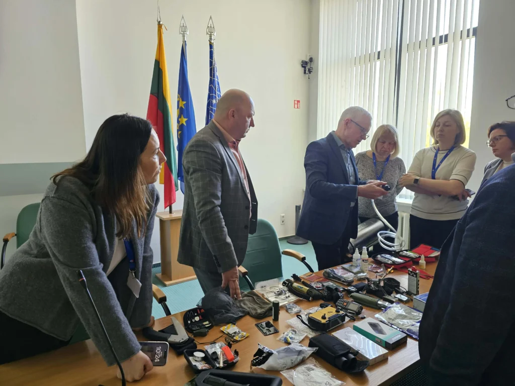 Asmenų grupė kabinete apžiūri ant stalo išdėliotą civilinės saugos įrangą ir pirmosios pagalbos priemones.