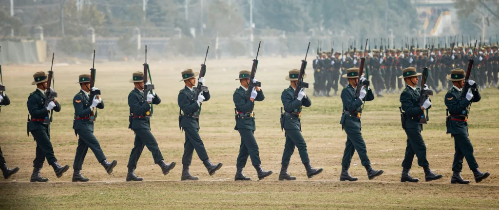 Kareiviai su žaliomis uniformomis ir skrybėlėmis žygiuoja lauke, laikydami šautuvus prie krūtinės iškilmingoje rikiuotėje.