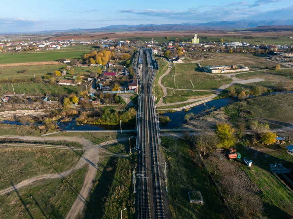 Geležinkelio bėgiai iš viršaus, einantys per tiltą virš upės į nedidelį miestelį kalvų fone.