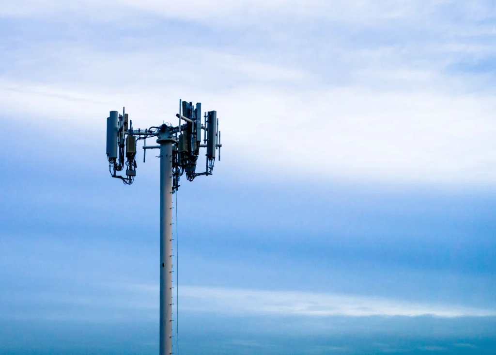 Aukštas pilkas telekomunikacijų bokštas su sumontuotomis antenomis šviesiai mėlyno, debesuoto dangaus fone.