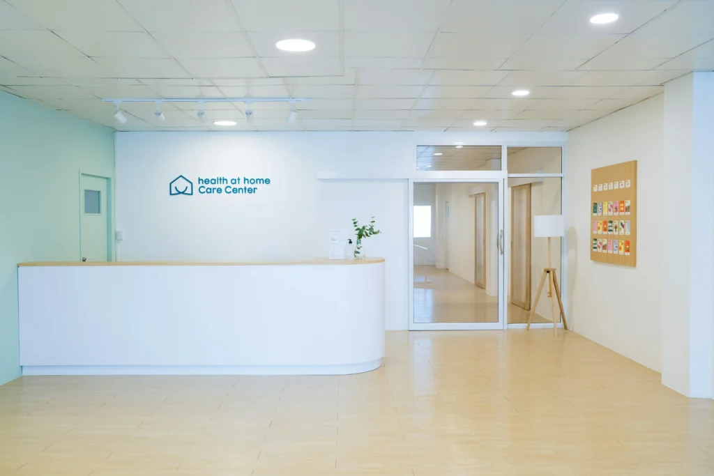 Moderni „Health at Home Care Center“ registratūra su baltu prekystaliu, šviesiomis grindimis ir minimalistiniu interjeru.