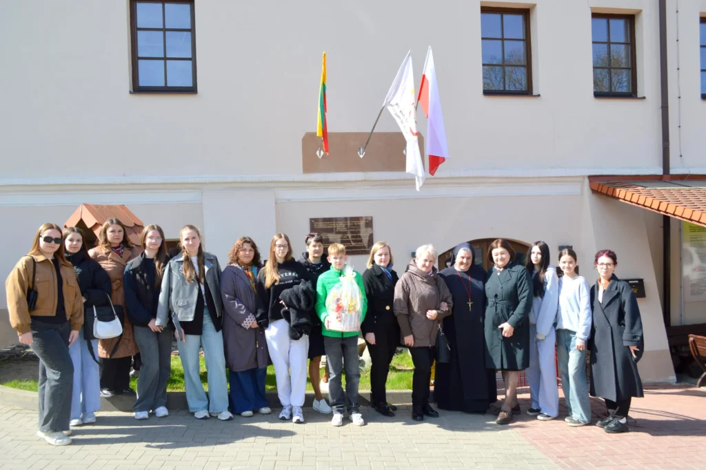 Grupė jaunimo ir moterų su vienuole stovi prie pastato su iškeltomis Lietuvos bei Lenkijos vėliavomis.