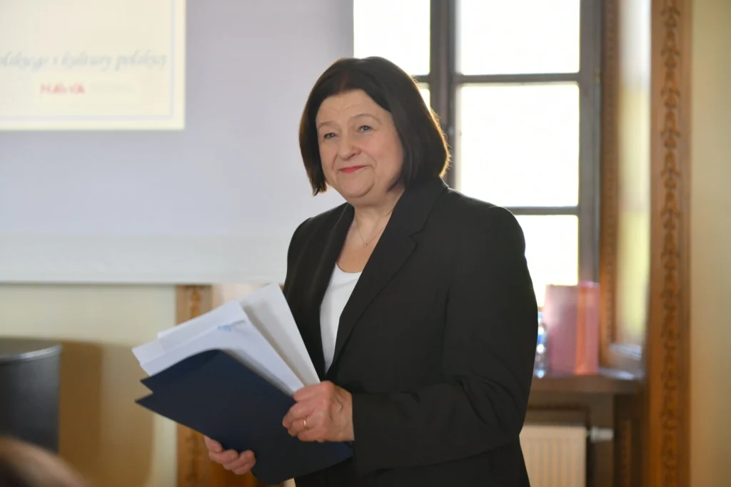 Švarku vilkinti tamsiaplaukė moteris šypsosi rankose laikydama popierius ir mėlyną dokumentų aplanką.