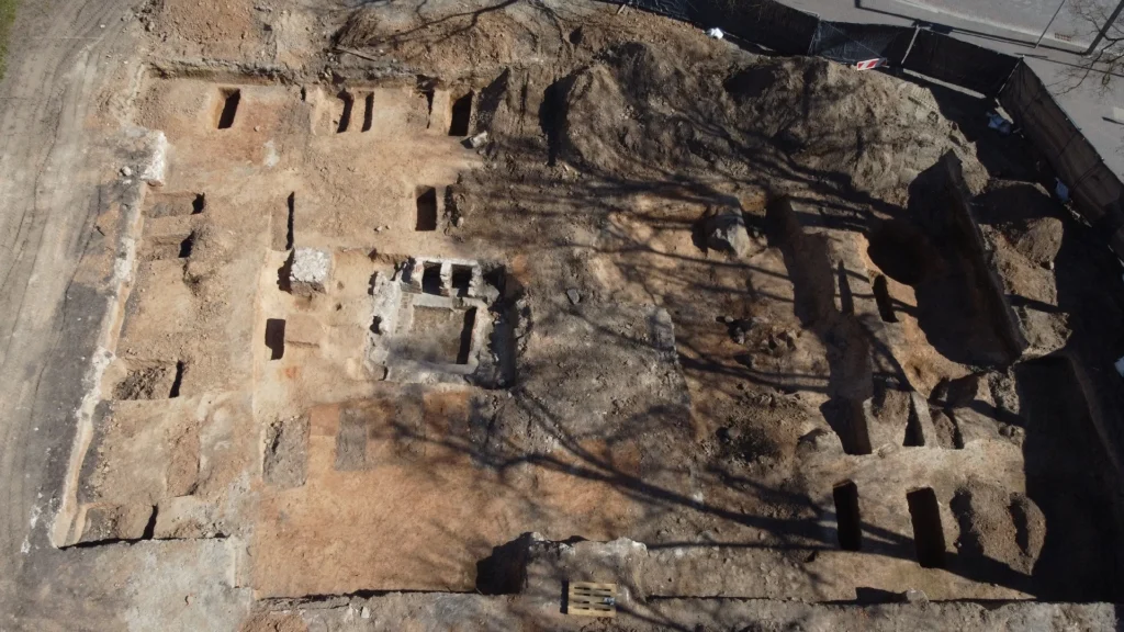 Iš viršaus matoma archeologinių kasinėjimų vieta su atviromis stačiakampėmis duobėmis ir senovinių pamatų liekanomis.