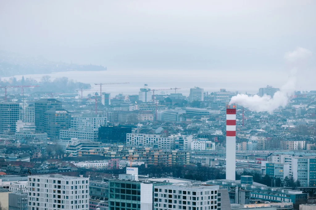 Miesto panorama su tankiais pastatais ir aukštu, raudonai baltu kaminu, iš kurio kyla balti dūmai.