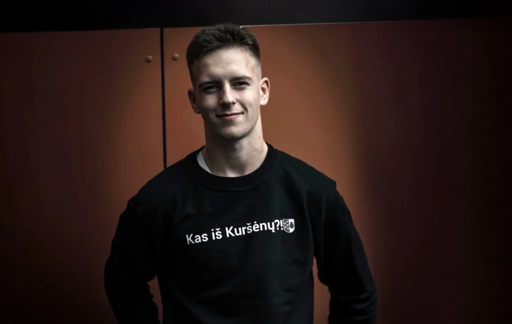 Besišypsantis jaunas vyras su juodais marškinėliais, papuoštais užrašu „Kas iš Kuršėnų?!“, rudame fone.