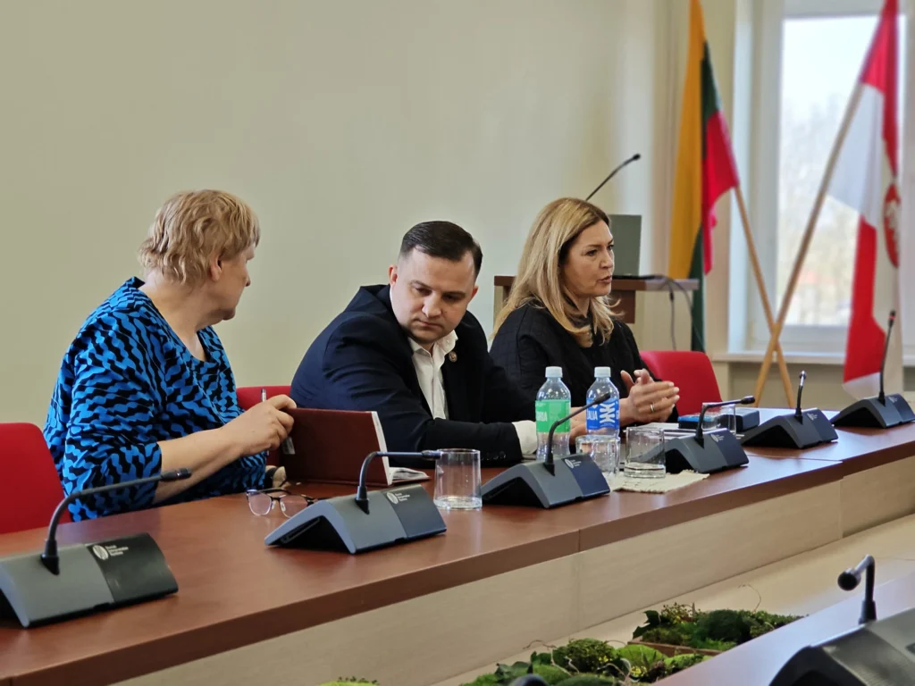 Trys asmenys sėdi prie posėdžių stalo su mikrofonais, fone matomos Lietuvos ir savivaldybės vėliavos.