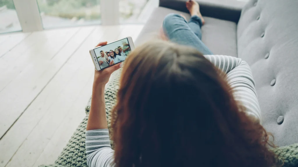 Moteris ant sofos per išmanųjį telefoną žiūri į besišypsančių draugų grupės nuotrauką šviesioje aplinkoje.