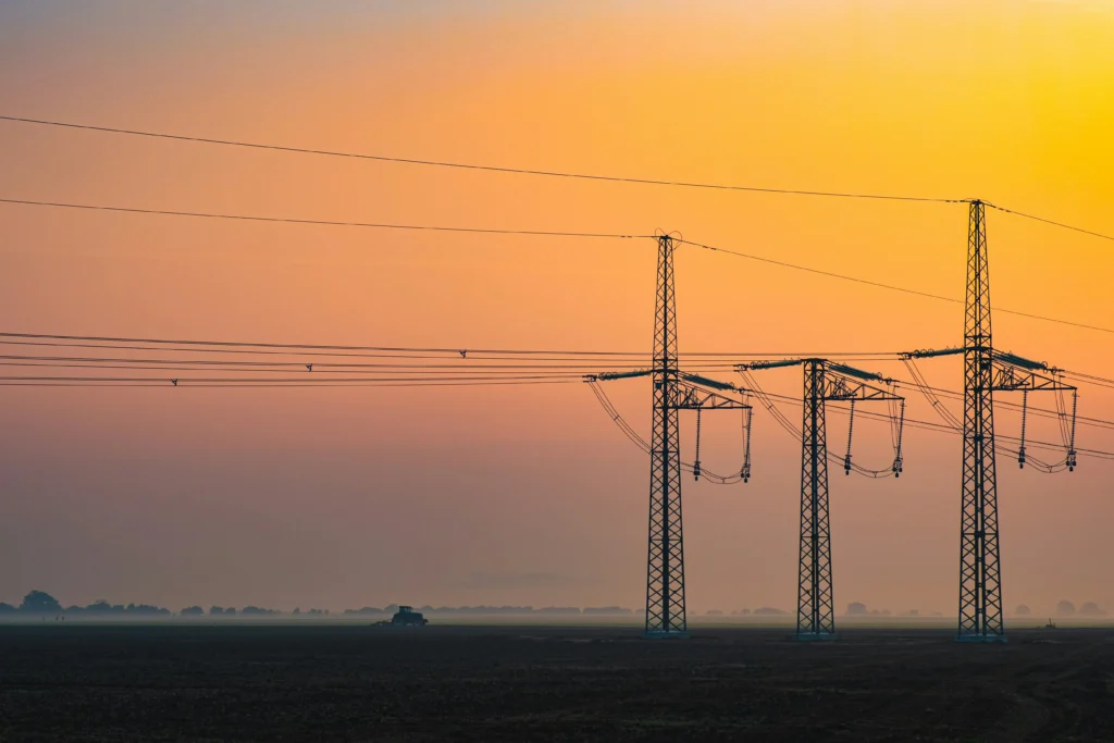 Trys aukštos įtampos elektros perdavimo bokštai tamsiame lauke po ryškiu oranžiniu saulėtekio dangumi.