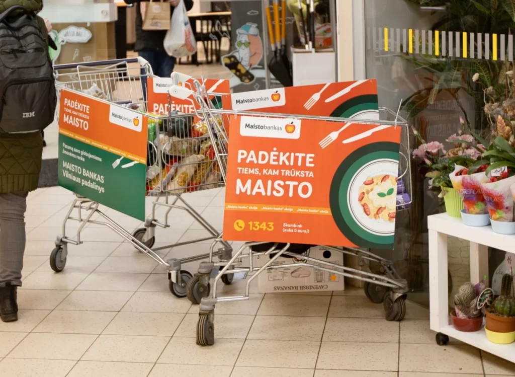 Parduotuvėje stovintys prekių vežimėliai su ryškiai oranžiniais „Maisto banko“ užrašais, kviečiančiais aukoti nepasiturintiems.
