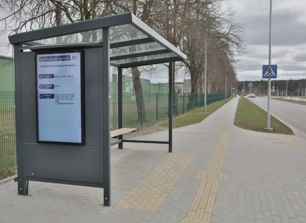 Moderni autobusų stotelė su skaitmeniniu ekranu, stikliniu stogu ir taktiliniu šaligatviu pilką rudens dieną.