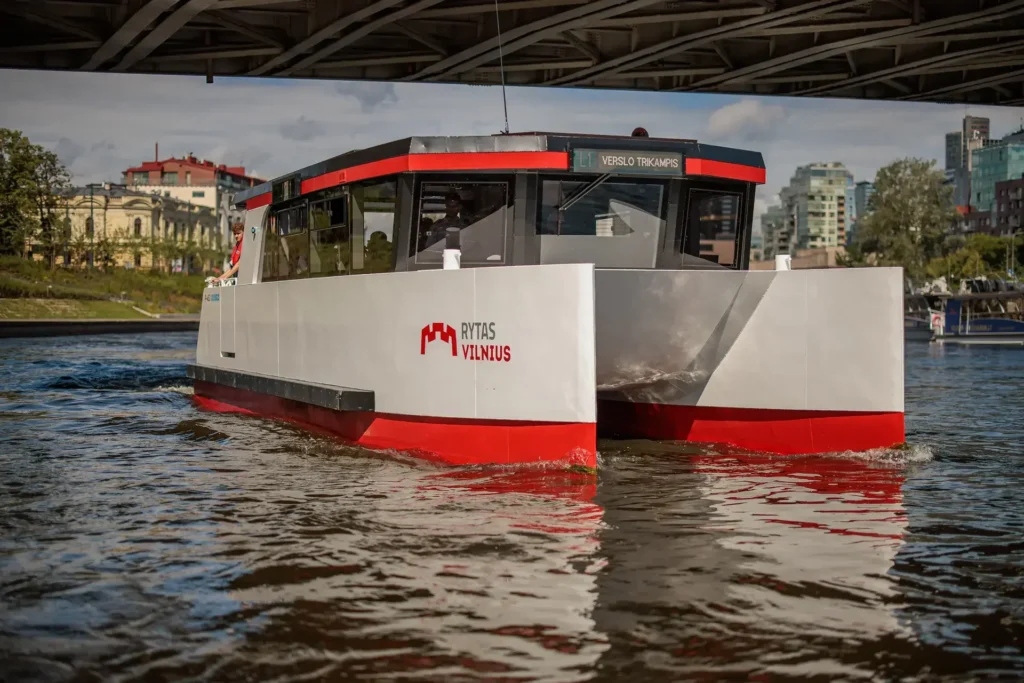 Modernus baltas ir raudonas keleivinis katamaranas su „Rytas“ simbolika plaukia Neries upe po tiltu Vilniuje.