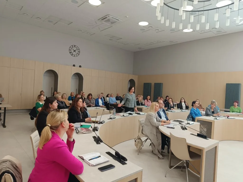 Pranešėja kalba auditorijai modernioje konferencijų salėje, kurioje dalyviai sėdi prie lenktų stalų po dideliu šviestuvu.
