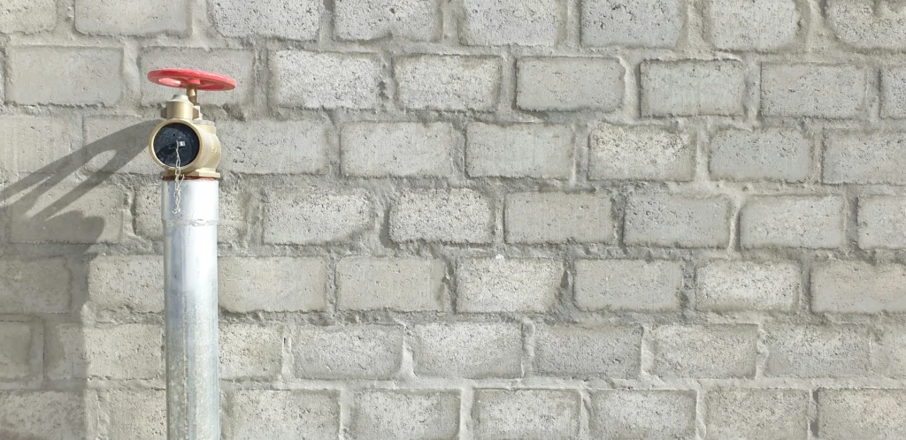 Prie pilkų betoninių blokelių sienos stovintis cinkuotas vamzdis su žalvarine jungtimi ir raudonu vožtuvu.