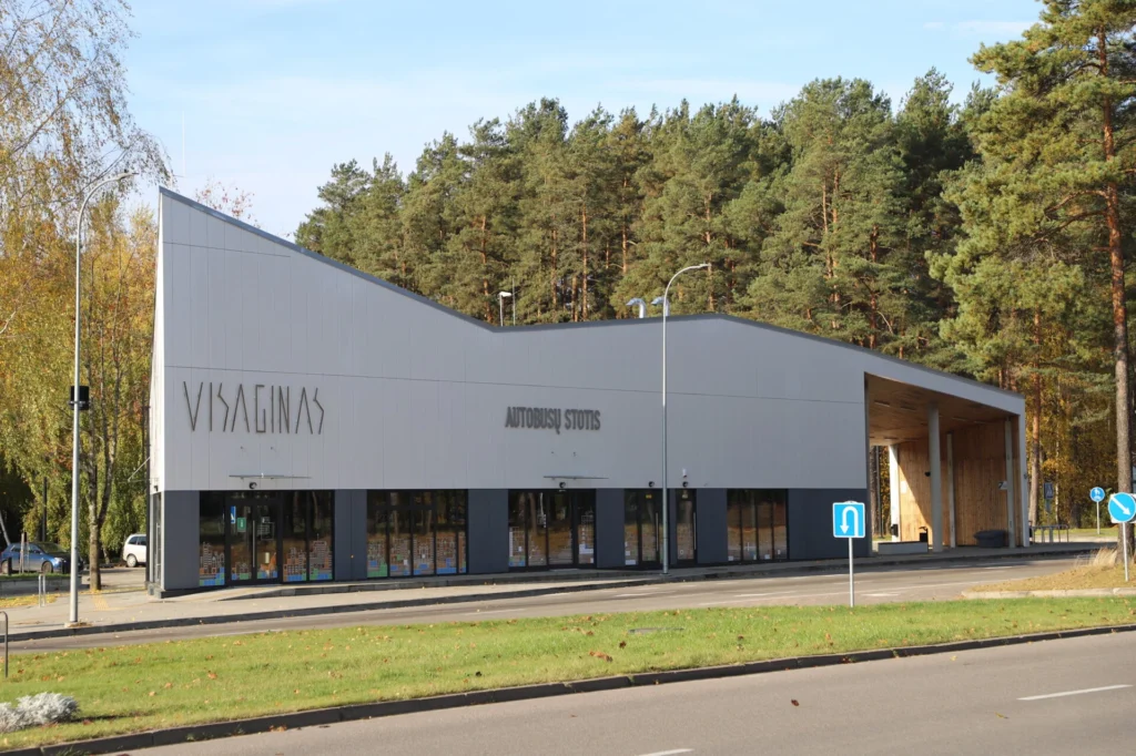 Modernus pilkšvas Visagino autobusų stoties pastatas su įstrižu stogu, stovintis šalia miško saulėtą dieną.