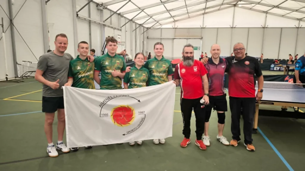 Septynių žmonių grupė sporto salėje laiko išskleistą Akmenės rajono sporto centro vėliavą prie teniso stalų.