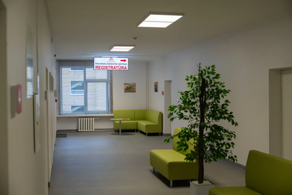 Šviesus medicinos įstaigos koridorius su žaliomis sofomis, augalu ir kabančia iškaba „Psichikos sveikatos centras Registratūra“.