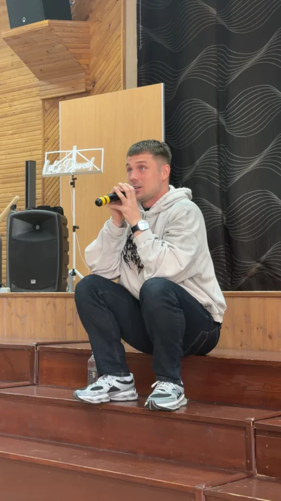 Vyras su šviesiu džemperiu pritūpęs ant medinių laiptų kalba į mikrofoną, fone matyti garso kolonėlė.