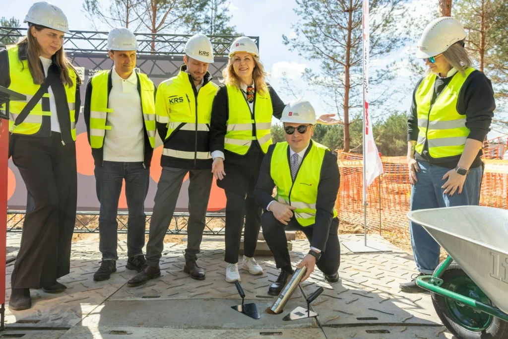 Grupė žmonių su apsauginėmis liemenėmis ir šalmais stebi vyrą, dedantį laiko kapsulę į pamatų angą.