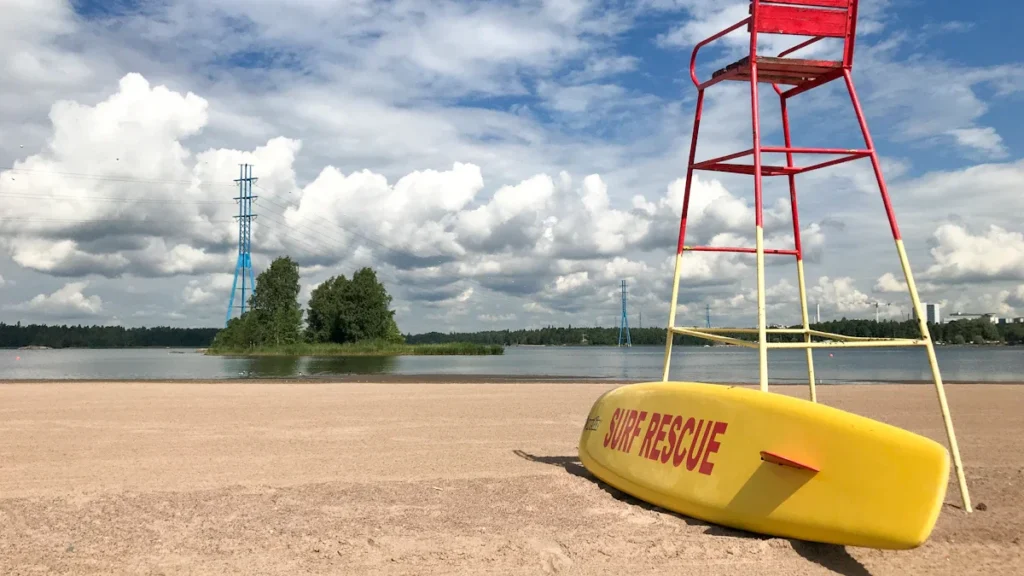Gelbėtojų stebėjimo bokštelis ir geltona gelbėjimo lenta tuščiame smėlio paplūdimyje prie ramaus vandens telkinio.