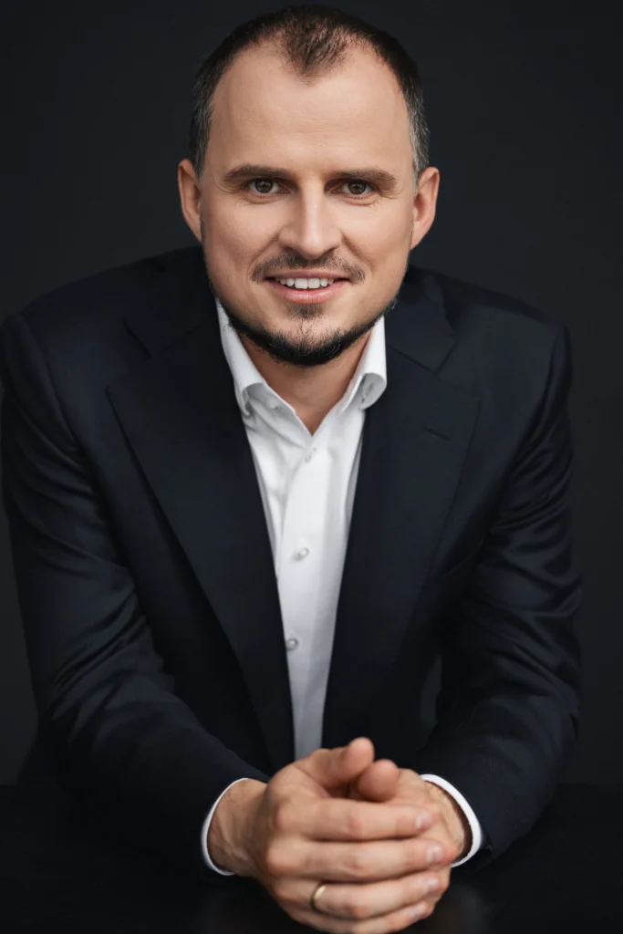Besišypsantis vyras su trumpa barzda, vilkintis juodą švarką, baltus marškinius ir sudėjęs rankas ant stalo.