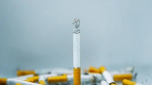 Viena degančia cigaretė tarp daugybės nuorūkų pilkame fone, simbolizuojanti priklausomybę.