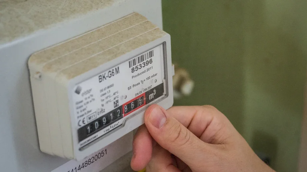 Žmogaus ranka liečia dujų skaitiklio rodmenis namų aplinkoje, iliustruojanti vartotojų energijos suvartojimo kontrolę.
