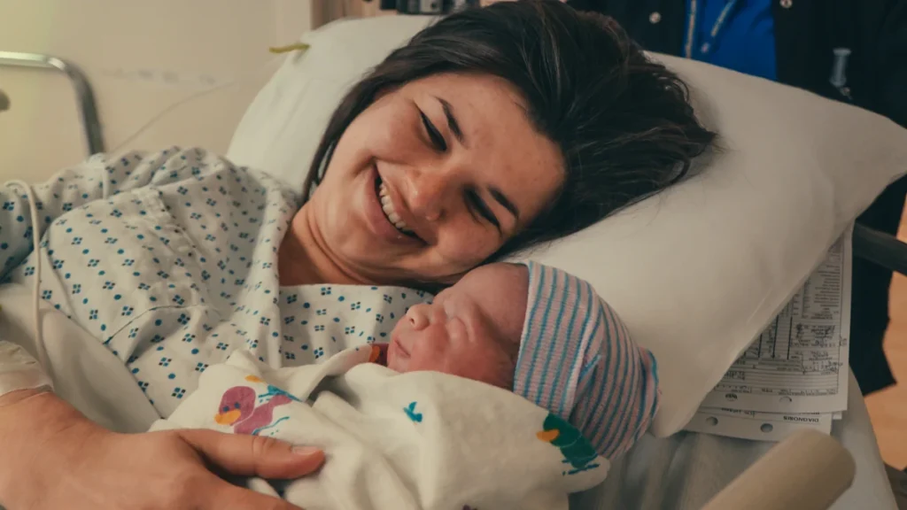 Jauna besišypsanti mama ligoninės lovoje švelniai glaudžia savo suvystytą naujagimį po sėkmingo gimdymo.