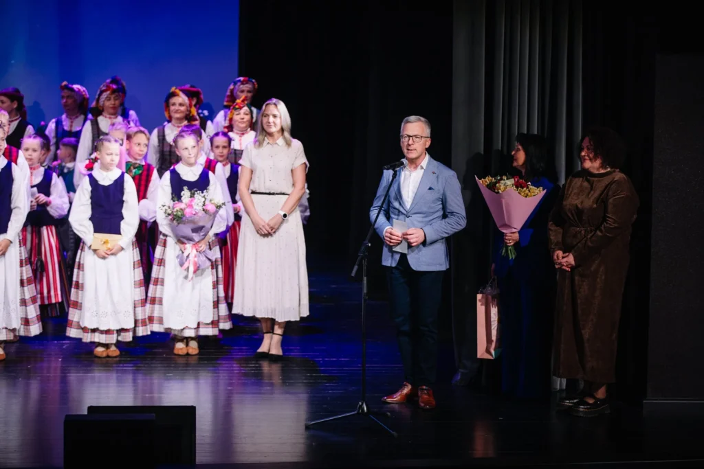 Vyras su švarku kalba į mikrofoną scenoje šalia vaikų grupės, vilkinčios tradicinius lietuviškus tautinius kostiumus.