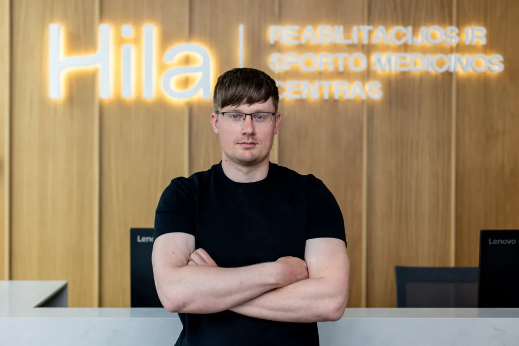 Vyras su akiniais ir juodais marškinėliais stovi sukryžiavęs rankas priešais „Hila“ medicinos centro šviečiantį logotipą.