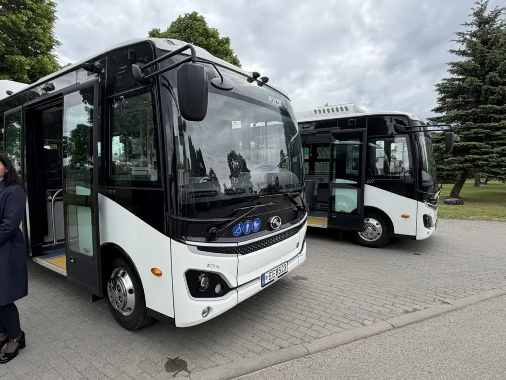 Du modernūs balti miesto autobusai stovi aikštelėje atviromis durimis debesuotą dieną.