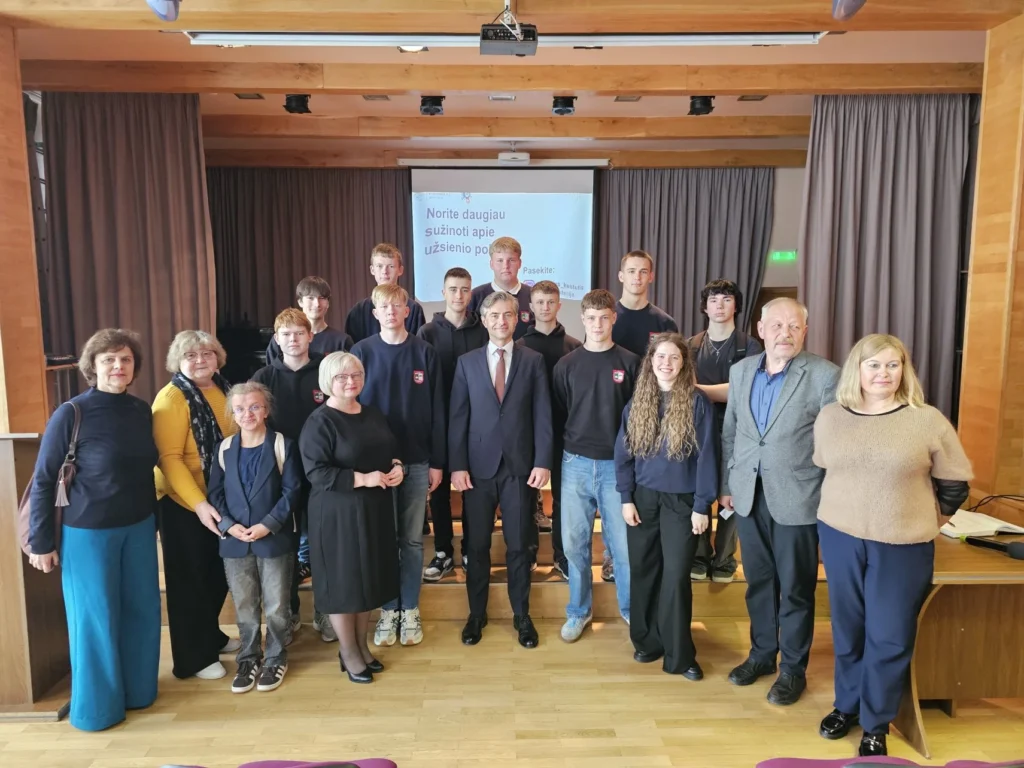 Didelė moksleivių ir suaugusiųjų grupė pozuoja nuotraukai ant scenos priešais projektoriaus ekraną.