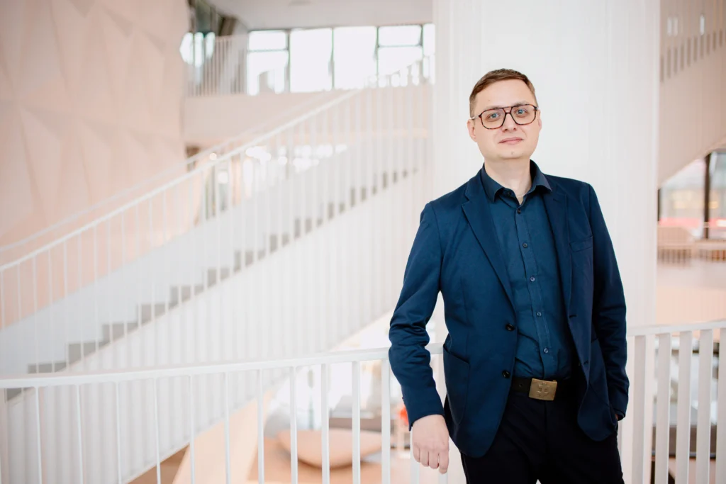 Vyras su akiniais ir tamsiai mėlynu švarku stovi prie baltų laiptų turėklų moderniame pastate.
