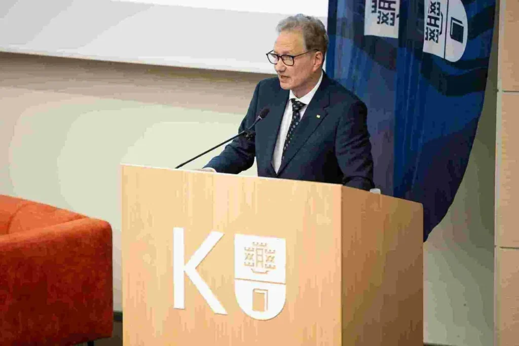 Vyresnio amžiaus vyras su akiniais ir kostiumu kalba prie universiteto tribūnos su logotipu.