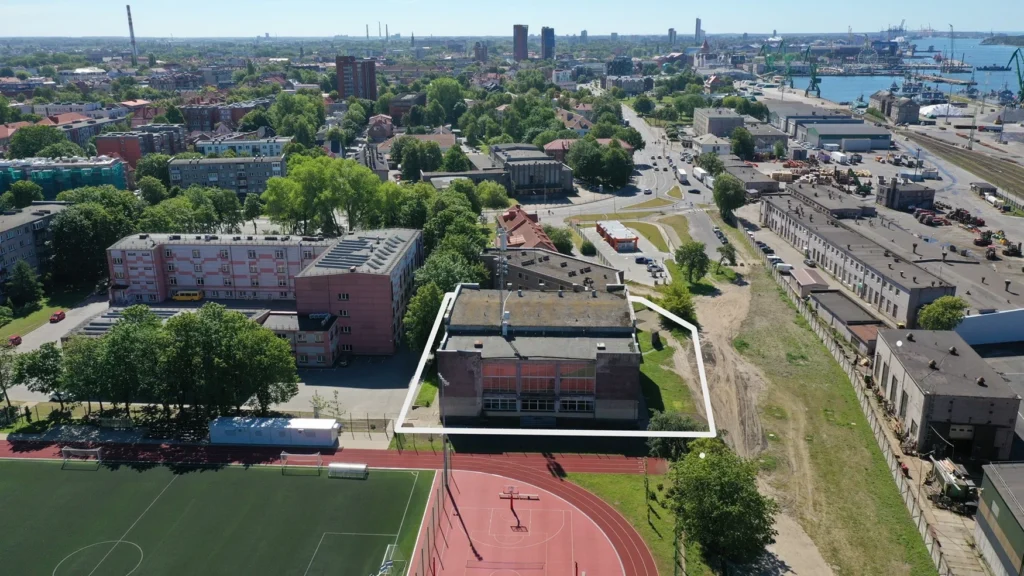 Miesto panorama iš paukščio skrydžio su balta linija apibrėžtu pastatu šalia stadiono ir uosto.