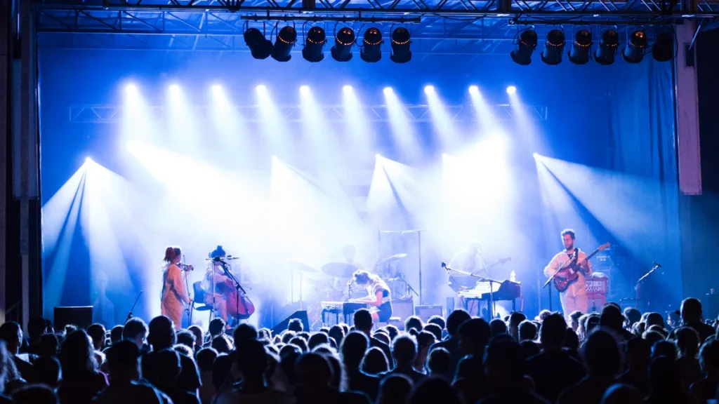 Muzikos grupė koncertuoja ryškiai apšviestoje scenoje priešais gausią žiūrovų minią vakarinio koncerto metu.