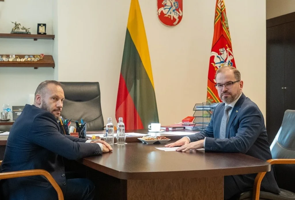 Du kostiumais vilkintys vyrai sėdi prie stalo, fone matomos Lietuvos valstybinė ir istorinė vėliavos.