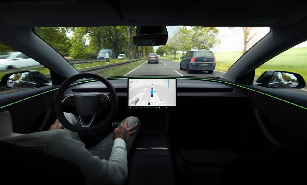 Tesla automobilio vidus, kuriame matomas ekranas su navigacija ir vairuotojas, nelaikantis vairo važiuojant keliu.